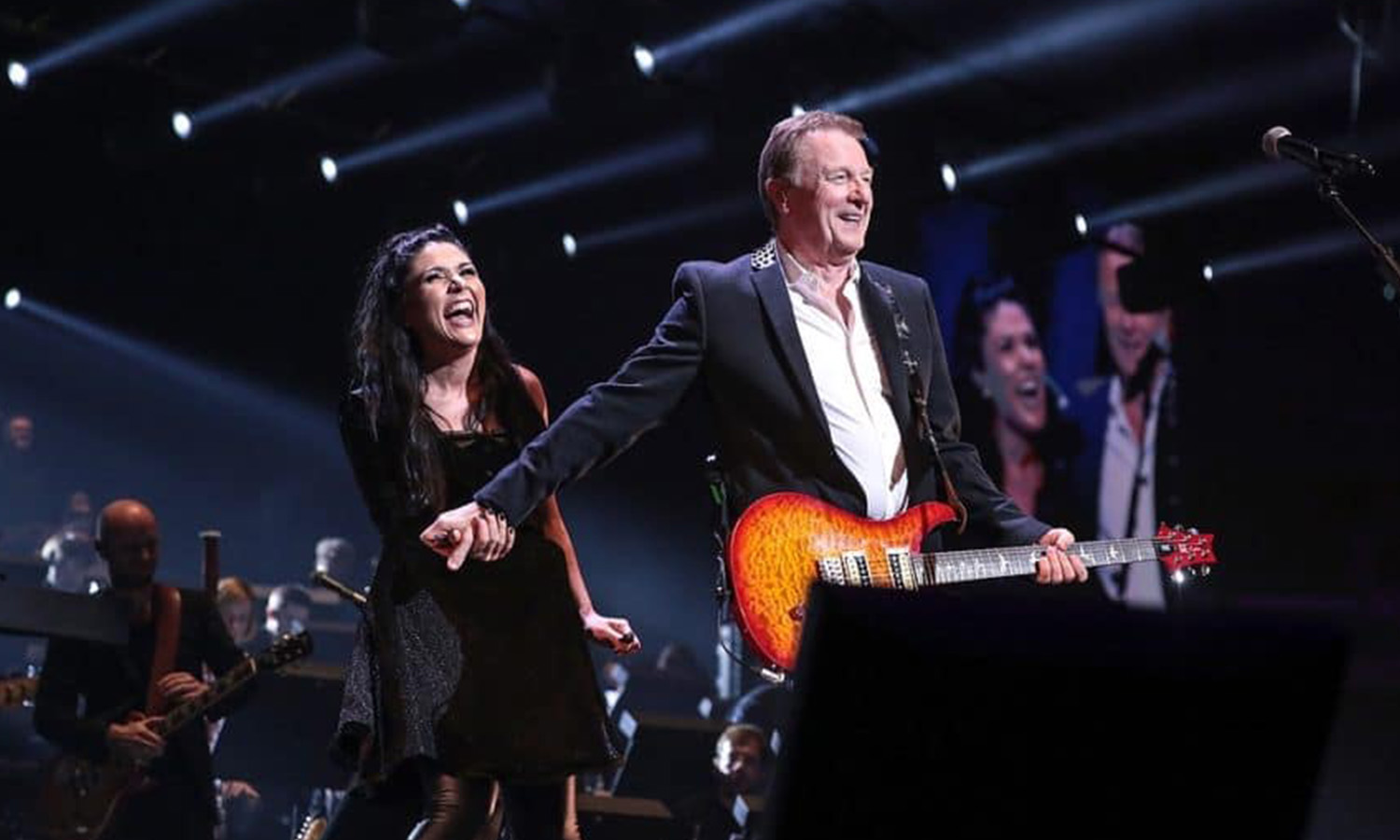Orchester und Künstler stehen gemeinsam auf der beleuchteten Bühne in der SAP Arena Mannheim während der Night of the Proms jubelnd vor Publikum.