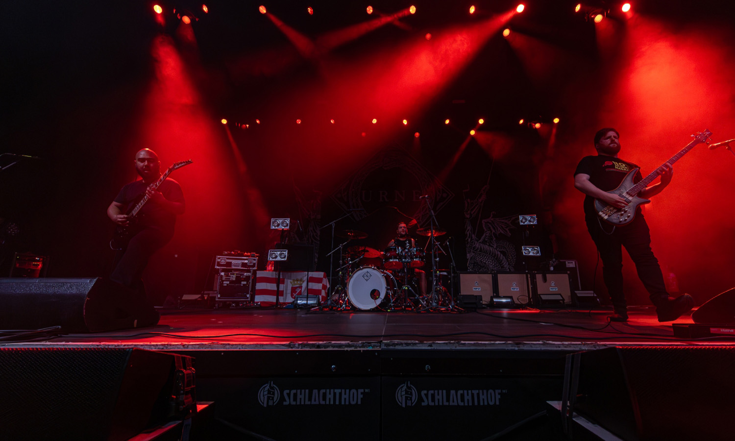 Eine Metal-Band performt energiegeladen auf der abgedunkelten Bühne des Schlachthofs Wiesbaden unter buntem Scheinwerferlicht, während die Menge feiert.
