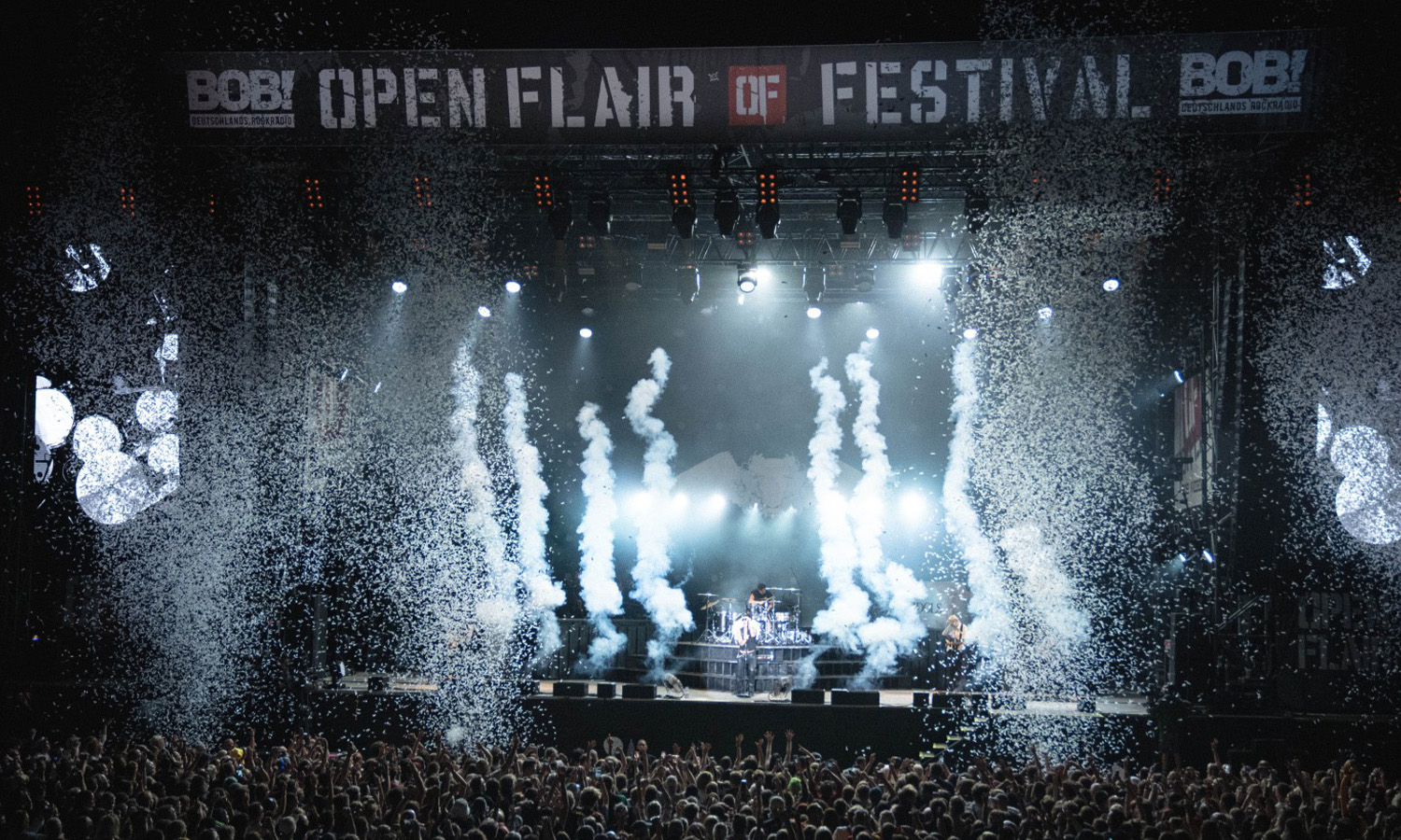 Eine ausgelassene Menge feiert vor einer großen Festivalbühne beim Open Flair in Eschwege, umgeben von bunten Lichtern und ausgelassener Stimmung.